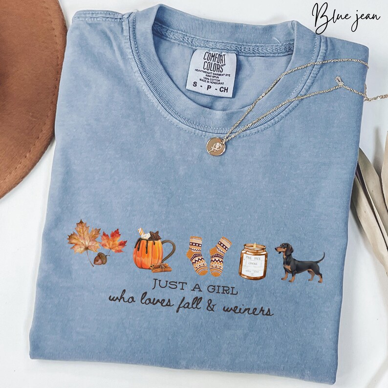 May include: Blue jean colored t-shirt with the text "JUST A GIRL who loves fall & weiners". The shirt features fall-themed graphics including leaves, a pumpkin spice latte, socks, a candle, and a dachshund dog.