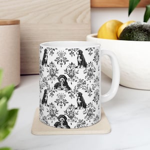 May include: White ceramic mug featuring a repeating pattern of black and white dog illustrations and floral damask designs. The mug has a curved handle and sits on a square coaster. The mug is a kitchen and dining accessory.