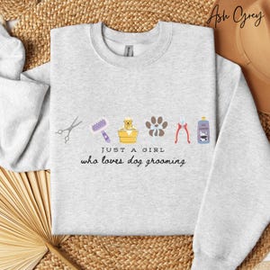 May include: Pale grey sweatshirt with dog grooming illustrations. Includes scissors, a brush, a dog in a tub, paw prints, clippers, and a shampoo bottle. Text reads "JUST A GIRL who loves dog grooming."