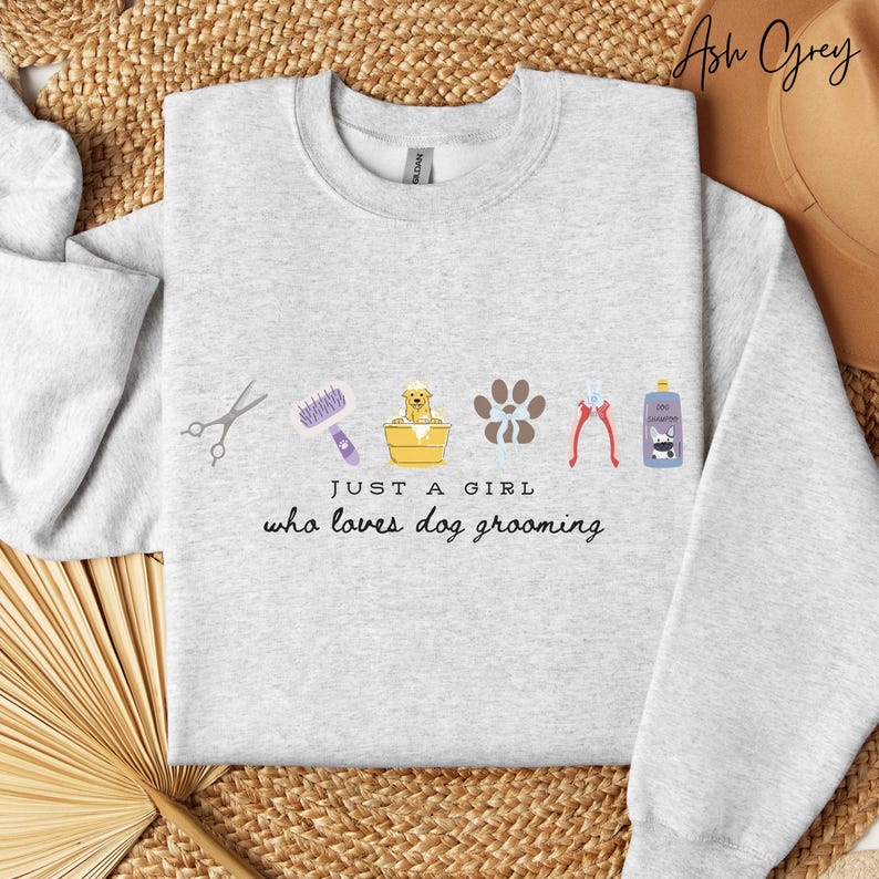 May include: Pale grey sweatshirt with dog grooming illustrations. Includes scissors, a brush, a dog in a tub, paw prints, clippers, and a shampoo bottle. Text reads "JUST A GIRL who loves dog grooming."