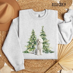May include: A white dog with a fluffy coat stands in front of two watercolor-style Christmas trees. The trees are green with white lights and the dog is looking at the camera. The text "ASH GREY" is in the top right corner of the image.