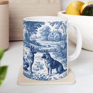 May include: A white ceramic mug with a blue and white design featuring two German Shepherd dogs in a pastoral setting. The design includes a house, trees, and flowers.