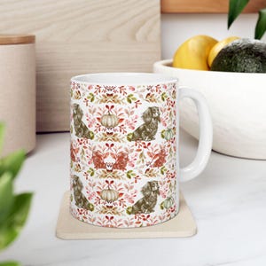 May include: White ceramic mug featuring a repeating pattern of dachshunds, pumpkins, and fall foliage in shades of brown, orange, and green. The mug has a curved handle and is placed on a beige coaster. The overall theme is autumn and the product is a mug.