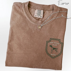 May include: Espresso-colored t-shirt with a crew neck and a small pocket design. The design features a green shield with the text "GSP DADS CLUB" and a dog illustration. A silver necklace with a pendant is draped across the shirt.