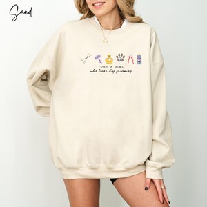 May include: A sand-coloured sweatshirt featuring the text "JUST A GIRL who loves dog grooming" and illustrations of grooming tools. The design includes scissors, a brush, a dog in a tub, a paw print, clippers, and a bottle.