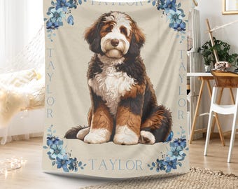 May include: A beige throw blanket featuring a seated Bernedoodle dog illustration. The dog has brown, black, and white fur. The name "TAYLOR" is printed in blue vertically on the sides and horizontally at the bottom, with blue floral accents.