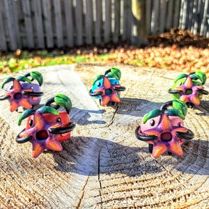 May include: A collection of six small, hand-painted figurines of a pink and green flower-like creature with black eyes. The figurines are arranged on a wooden surface.