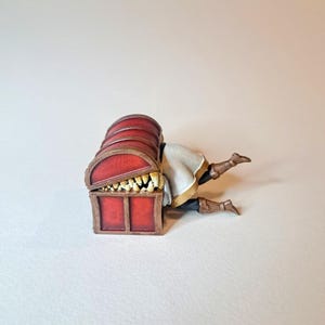 May include: A red wooden treasure chest with a hinged lid is open, revealing a set of sharp teeth inside. A miniature figure of a person is trapped inside the chest, with their legs sticking out.
