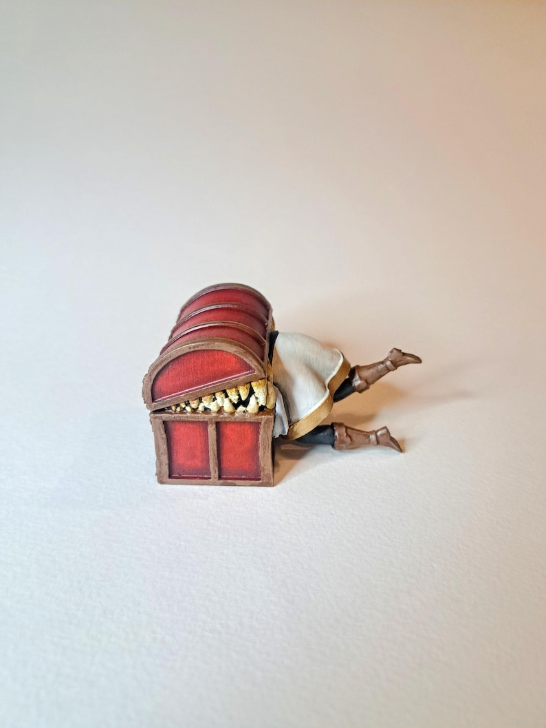 May include: A red wooden treasure chest with a hinged lid is open, revealing a set of sharp teeth inside. A miniature figure of a person is trapped inside the chest, with their legs sticking out.