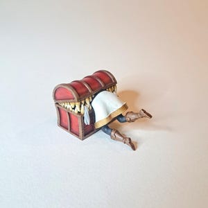 May include: A miniature figurine of a person trapped inside a closed wooden treasure chest with gold coins inside. The chest is red with gold accents. The person is wearing a white tunic and brown boots.