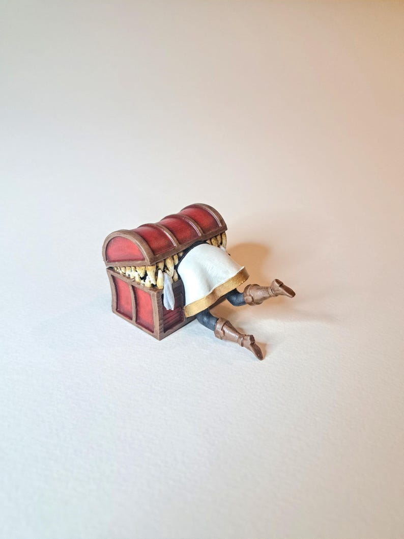 May include: A miniature figurine of a person trapped inside a closed wooden treasure chest with gold coins inside. The chest is red with gold accents. The person is wearing a white tunic and brown boots.