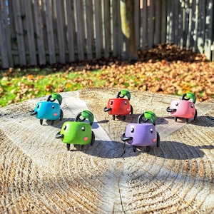 May include: Five small, colourful, hand-painted figurines with a cartoon-like design. Each figurine has a green leaf on top and a black outline. The figurines are arranged on a wooden surface.