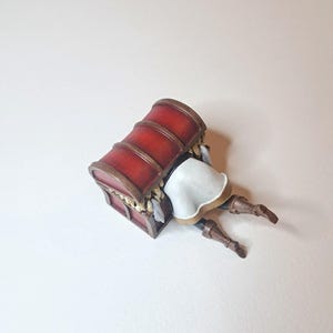 May include: A red wooden treasure chest with gold accents is lying on its side. A figurine of a person is peeking out from the chest, with their legs sticking out.