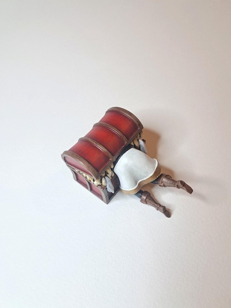 May include: A red wooden treasure chest with gold accents is lying on its side. A figurine of a person is peeking out from the chest, with their legs sticking out.