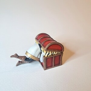 May include: A red wooden treasure chest with gold accents and a white fabric lining. A miniature figure is hiding inside the chest.