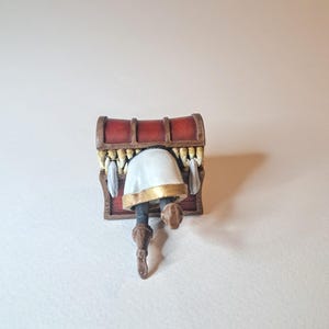 May include: A small, red wooden treasure chest with a white and gold figure inside. The figure is upside down with its legs sticking out of the chest.