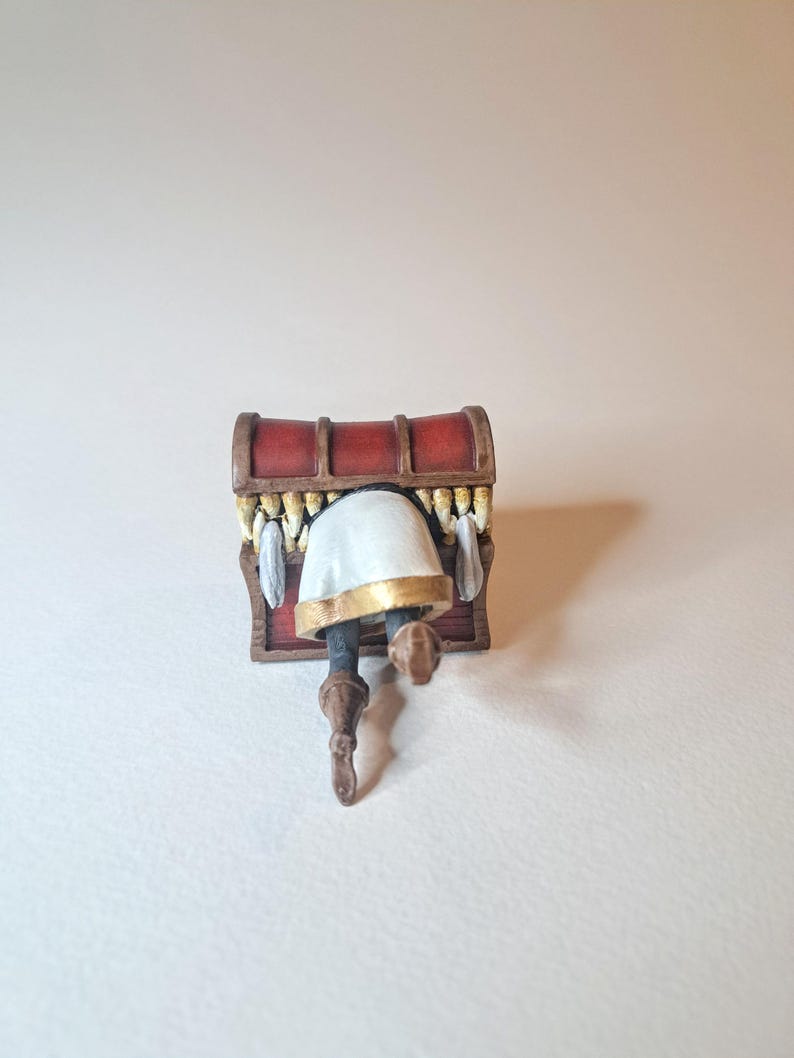 May include: A small, red wooden treasure chest with a white and gold figure inside. The figure is upside down with its legs sticking out of the chest.