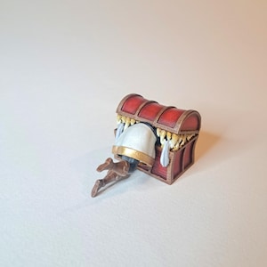May include: A red wooden treasure chest with gold accents is open and a miniature figure is partially inside. The figure is wearing a white tunic with gold trim and brown boots.