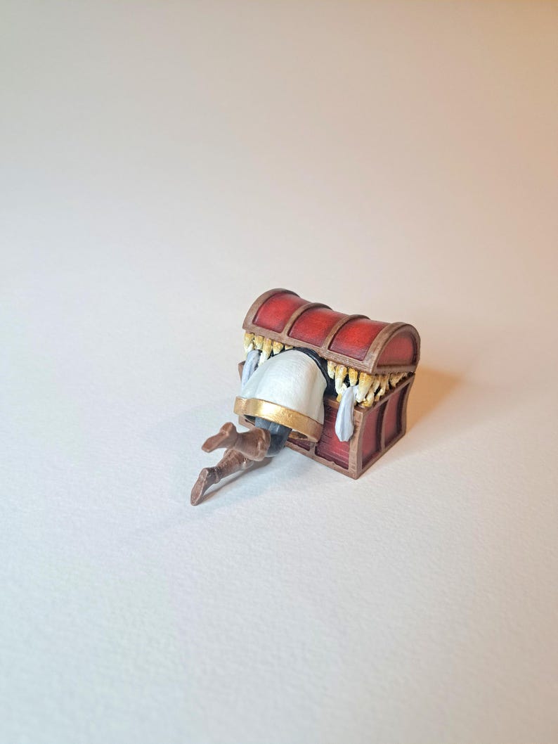 May include: A red wooden treasure chest with gold accents is open and a miniature figure is partially inside. The figure is wearing a white tunic with gold trim and brown boots.