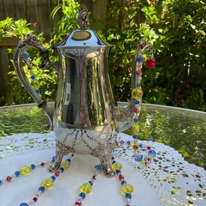 May include: A silver teapot with a beaded chain made of yellow, blue, red, and clear beads. The chain is draped over the teapot and hangs down to the table.