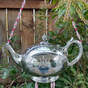 May include: A silver teapot wind chime with pink and clear glass beads hanging from it. The teapot is hanging from a chain of pink and clear glass beads.
