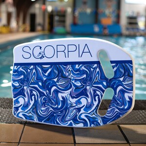 May include: A white and blue Scorpa kickboard with a swirling pattern. The kickboard has a handle and a light blue grip. The word "SCORPIA" is printed in blue on the top of the kickboard. The kickboard is next to a swimming pool.