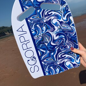 May include: A white and blue bodyboard with a swirling pattern of blues and purples. The board has two hand cutouts and the word "SORPIA" printed vertically in white with a blue border. The board is held on a sandy beach.