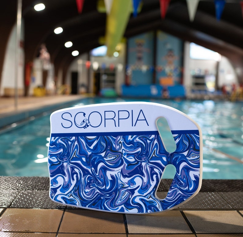 May include: A white and blue Scorpa kickboard with a swirling pattern. The kickboard has a handle and a light blue grip. The word "SCORPIA" is printed in blue on the top of the kickboard. The kickboard is next to a swimming pool.