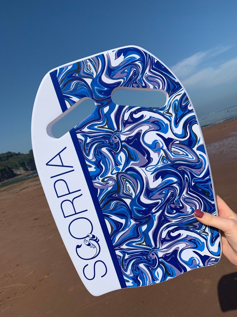 May include: A white and blue bodyboard with a swirling pattern of blues and purples. The board has two hand cutouts and the word "SORPIA" printed vertically in white with a blue border. The board is held on a sandy beach.