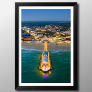 Può includere: Una vista aerea di un molo che si estende nell'oceano al tramonto. Il molo è illuminato da luci gialle e ci sono persone che camminano su di esso. Lo skyline della città è visibile sullo sfondo e l'acqua è di un blu scuro.