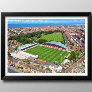 Könnte beinhalten: Luftaufnahme eines Fußballstadions mit einem grünen Rasen und roten und weißen Tribünen. Das Stadion ist von einem Wohngebiet und einem Gewässer in der Ferne umgeben.