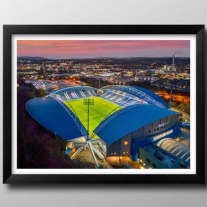 Könnte beinhalten: Gerahmte Luftaufnahme eines Stadions in der Abenddämmerung. Das Stadion hat ein blaues Dach und einen grünen Rasen, der von hellen Lichtern beleuchtet wird. Der Himmel ist eine Mischung aus Orange- und Rosatönen, und die Lichter der Stadt funkeln im Hintergrund.