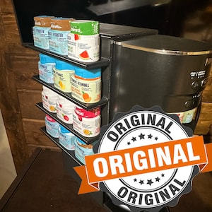 Può includere: Un distributore di bevande nero e argento con un contenitore di plastica trasparente per contenere acqua. Il distributore ha uno scaffale con diversi piccoli pacchetti colorati di miscela per bevande. I pacchetti sono etichettati con le parole "First" e "Vitamin C".