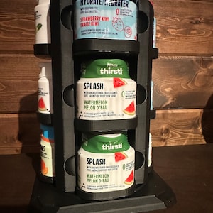 May include: A black rotating stand with three tiers holds six bottles of flavored water drops. The bottles are labeled with the brand "Ninja Thirst" and feature flavors like "Strawberry Kiwi" and "Watermelon Melon d'Eau".