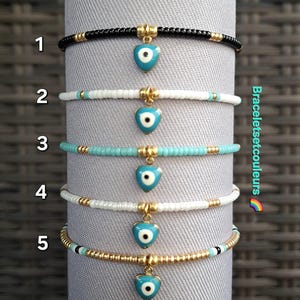 Peut inclure: Cinq bracelets de perles de couleurs variées : noir, blanc et turquoise. Chaque bracelet est orné d'un charm doré en forme de cœur avec un œil bleu. Les bracelets sont présentés sur une surface cylindrique grise.