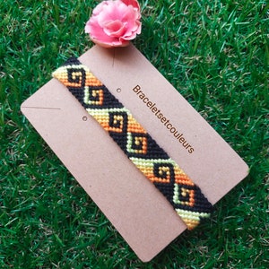 Op de afbeelding: Een zwart, oranje en geel vriendschapsarmband met een geometrisch patroon. De armband ligt op een bruine kaart met de tekst "Braceletsetcouleurs".