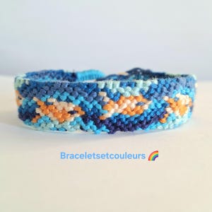 Op de afbeelding: Handgemaakte vriendschapsarmband in blauwe, oranje en witte tinten. Het geweven ontwerp heeft een herhalend patroon. De tekst "Braceletsetcouleurs" staat eronder, samen met een regenboogafbeelding.
