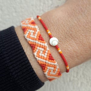 Op de afbeelding: Twee armbanden om een pols. De bovenste armband is een geweven vriendschapsarmband met een geometrisch patroon in oranje en wit. De onderste armband is een kralenarmband met rode en witte kralen en een witte schelpbedel.