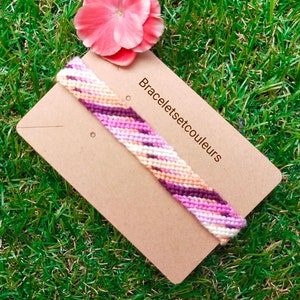 Op de afbeelding: Een roze, witte en paarse vriendschapsarmband met een chevron-patroon op een bruine kaart met de tekst "Braceletsetcouleurs".