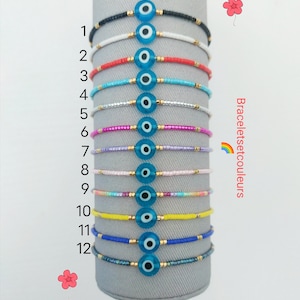 Peut inclure: Douze bracelets en perles colorées avec un charme œil bleu maléfique. Les bracelets sont disposés verticalement sur un présentoir gris. Les chiffres 1 à 12 sont imprimés à gauche des bracelets.