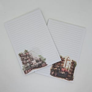 Könnte beinhalten: Zwei linierte Notizblöcke mit Aquarellillustrationen von gemütlichen Leseecken. Ein Notizblock zeigt ein Bücherregal und ein Sofa, der andere einen Lesesessel und weitere Bücherregale. Die Notizblöcke sind weiß mit grauen Linien.