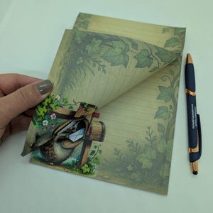 May include: Stationery set featuring lined paper with a decorative ivy border and a whimsical illustration of a hedgehog delivering mail. A blue pen with gold accents rests beside the paper. The paper has a vintage aesthetic.