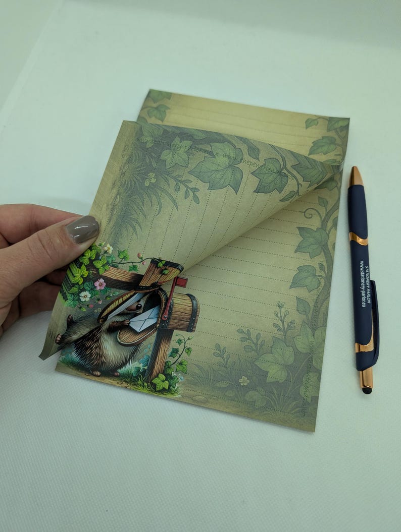 May include: Stationery set featuring lined paper with a decorative ivy border and a whimsical illustration of a hedgehog delivering mail. A blue pen with gold accents rests beside the paper. The paper has a vintage aesthetic.