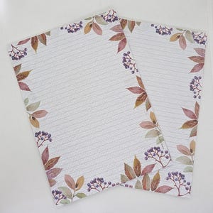 Può includere: Due fogli di carta da lettere a righe con un bordo decorativo di foglie autunnali in stile acquerello nei toni del marrone, beige e verde, intervallate da grappoli di piccole bacche viola scuro su uno sfondo bianco sporco.