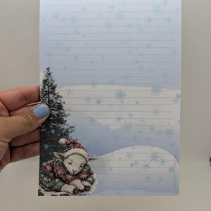 May include: Winter-themed stationery featuring a sleeping lamb wearing a red and green sweater and hat. The stationery has a light blue snowflake design and lined paper. A small evergreen tree is on the left side of the stationery.