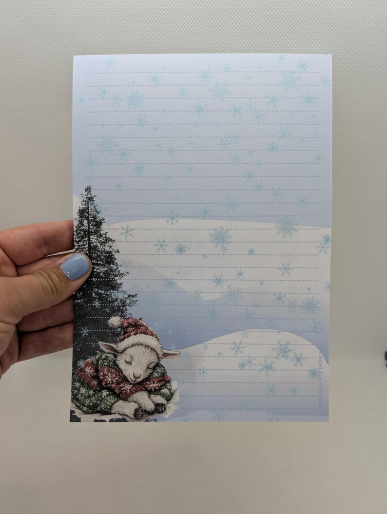 May include: Winter-themed stationery featuring a sleeping lamb wearing a red and green sweater and hat. The stationery has a light blue snowflake design and lined paper. A small evergreen tree is on the left side of the stationery.