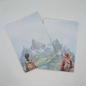 May include: Two lined notepads featuring a mountain landscape scene. The illustration depicts two people with backpacks gazing at a mountain range. The notepads have a light blue background with horizontal lines.