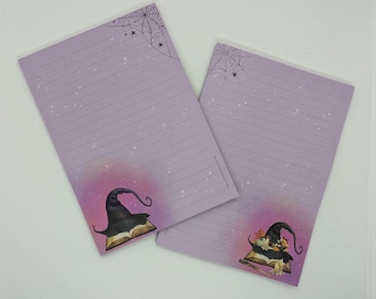 May include: Two purple lined stationery notepads with a Halloween theme. One notepad features a witch's hat on top of a book with a spider web in the corner. The other notepad features a witch with a wand and a book with a spider web in the corner.