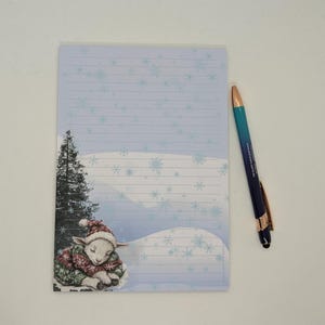 May include: A lined notepad with a winter scene featuring a sleeping lamb in a Christmas sweater and hat. A blue and gold pen rests beside the notepad. The notepad has a light blue background with snowflake designs.