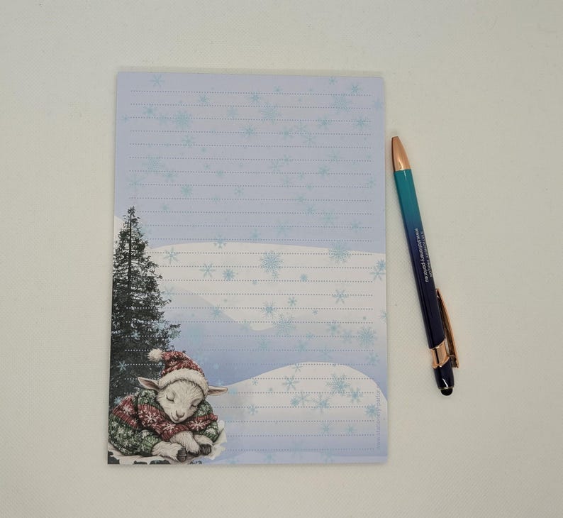 May include: A lined notepad with a winter scene featuring a sleeping lamb in a Christmas sweater and hat. A blue and gold pen rests beside the notepad. The notepad has a light blue background with snowflake designs.
